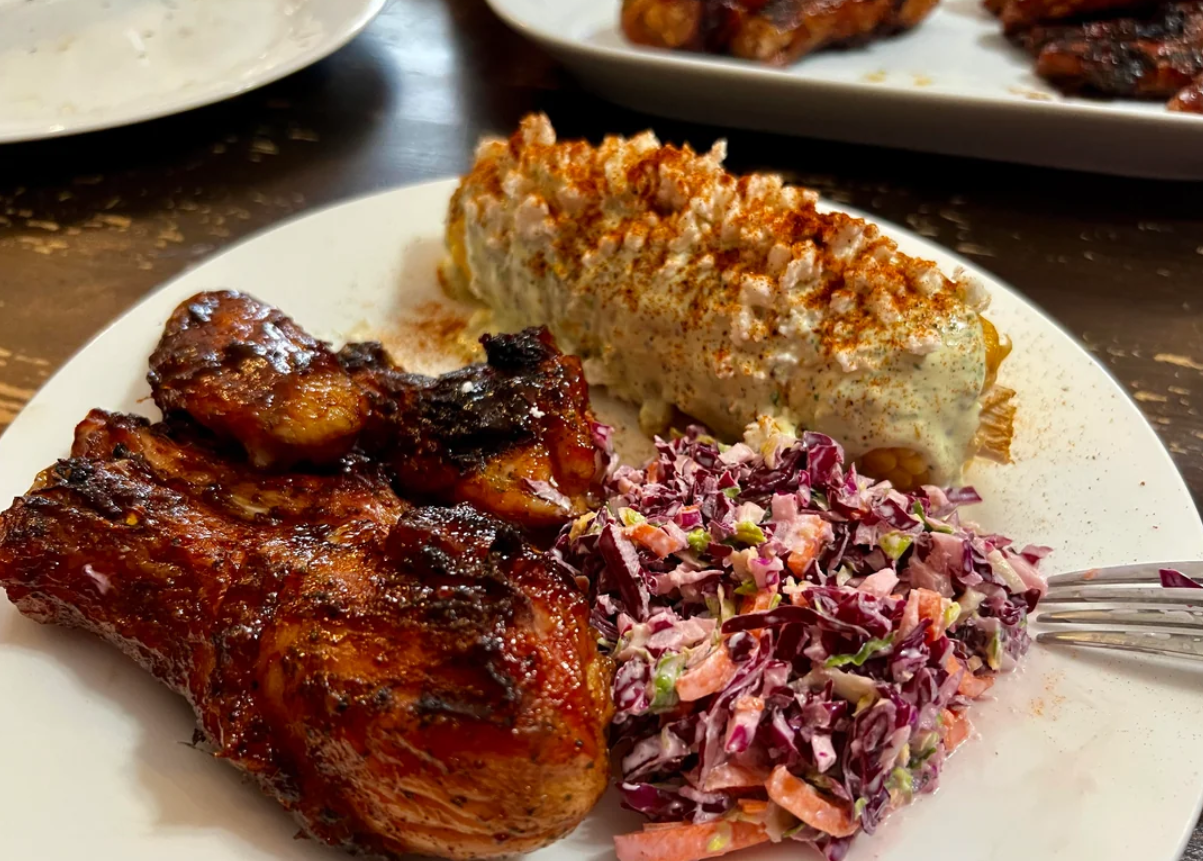 A plate with barbecued chicken, elote topped with cheese and spices, and a serving of colorful coleslaw. A fork is next to the food.