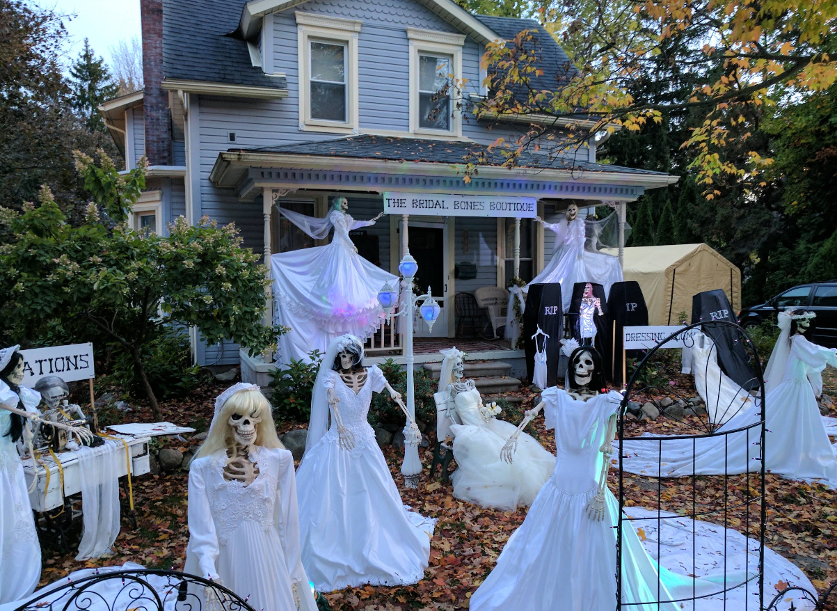 A house decorated for Halloween with skeletons in white dresses, ghost figures, tombstones, and a sign reading "The Bridal Bones Boutique" in the yard captures the festive spirit found in some of the best trick or treat towns in America. Autumn leaves surround the scene.
