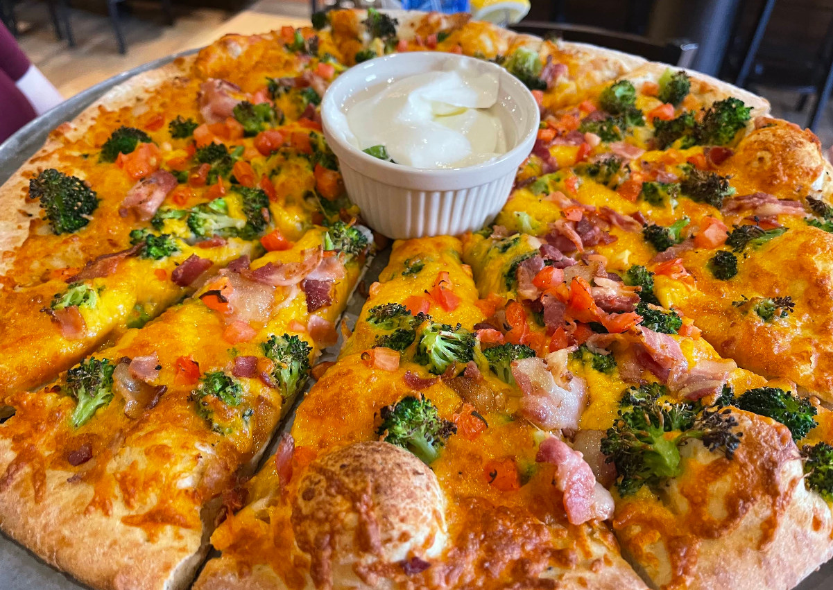 A close-up of a pizza topped with broccoli, diced tomatoes, bacon, and cheddar cheese—sliced and served with a cup of white dipping sauce in the center—offers a quality slice of pizza for cheap.