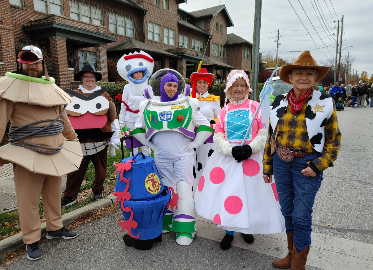 A group of adults in colorful Toy Story costumes, including Woody, Buzz Lightyear, Bo Peep, Forky, and others, pose together on a suburban street with brick houses in the background—capturing the festive spirit you’ll find in some of the best trick or treat towns in America.