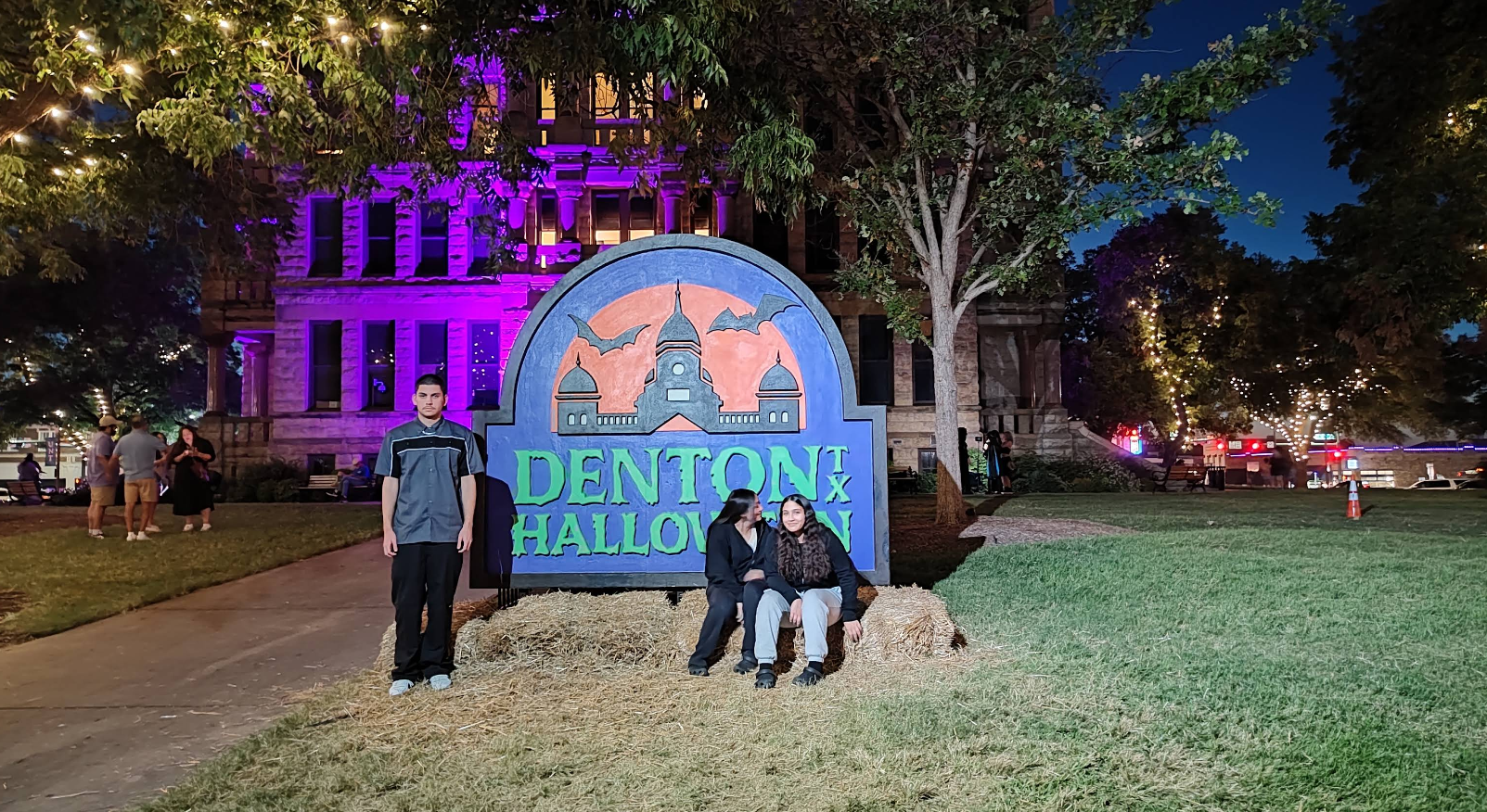 Three people pose by a large "Denton, TX Halloween" sign in front of a lit-up building at night, with purple and orange lights decorating the scene. Hay bales and trees surround the area, capturing the festive spirit that makes Denton one of the best trick or treat towns in America.