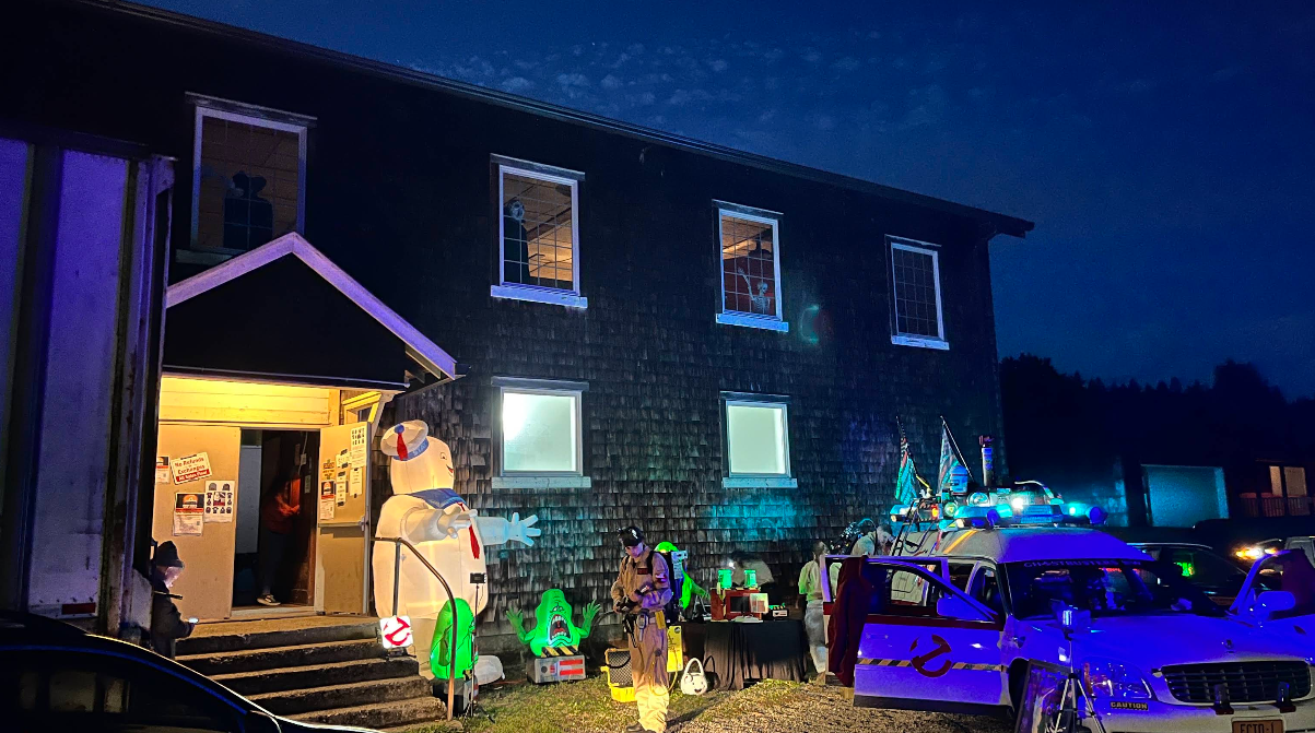 A building at night with Ghostbusters decorations, glowing green lights, and a car with blue lights on. People in costumes and props are outside the entrance, with an inflatable Stay Puft Marshmallow Man near the door—capturing the festive spirit found in some of the best trick or treat towns in America.