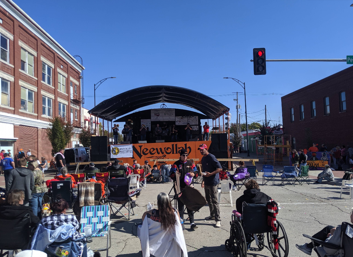 A crowd watches a band perform on an outdoor stage at a street festival called Neewollah, held in one of the best trick or treat towns in America. People sit in lawn chairs under a sunny sky, with buildings and a traffic light visible in the background.