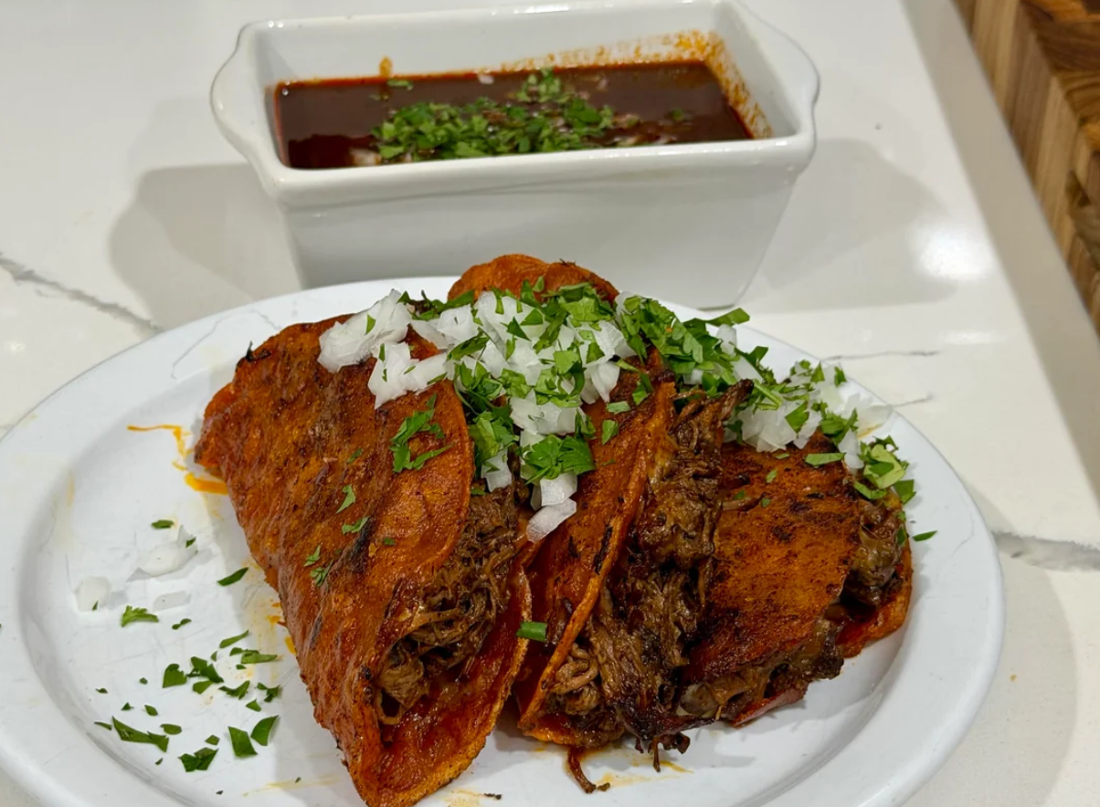 Two birria tacos filled with shredded meat, topped with chopped onions and cilantro, are served on a white plate. A bowl of rich, red consommé garnished with herbs sits behind them on a white countertop.