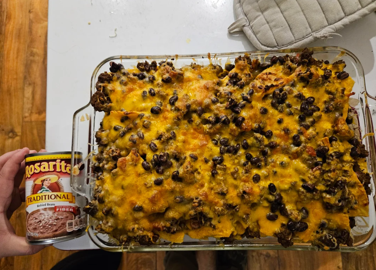 A glass baking dish filled with a cheesy casserole topped with black beans and melted cheese is on a table. A hand holds a can of Rosarita Traditional Refried Beans next to the dish. An oven mitt is nearby.