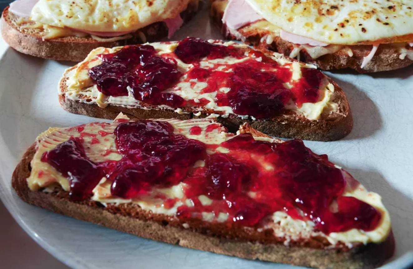 Two slices of bread covered with butter and raspberry jam on a plate, with additional open-faced sandwiches featuring eggs, ham, and cheese in the background.