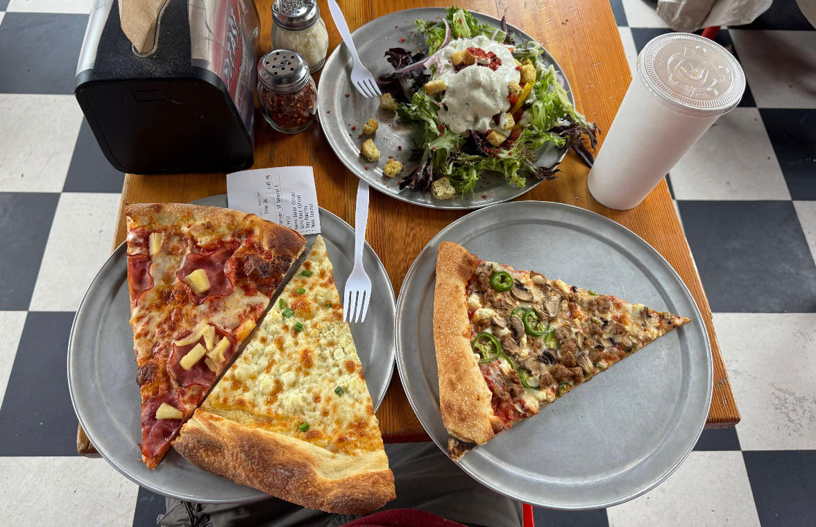 Two metal plates with large, quality slices of pizza for cheap—one topped with ham and pineapple, the other with mushrooms and jalapeños—sit on a wooden table alongside salad, a plastic cup, napkin holder, and condiments.