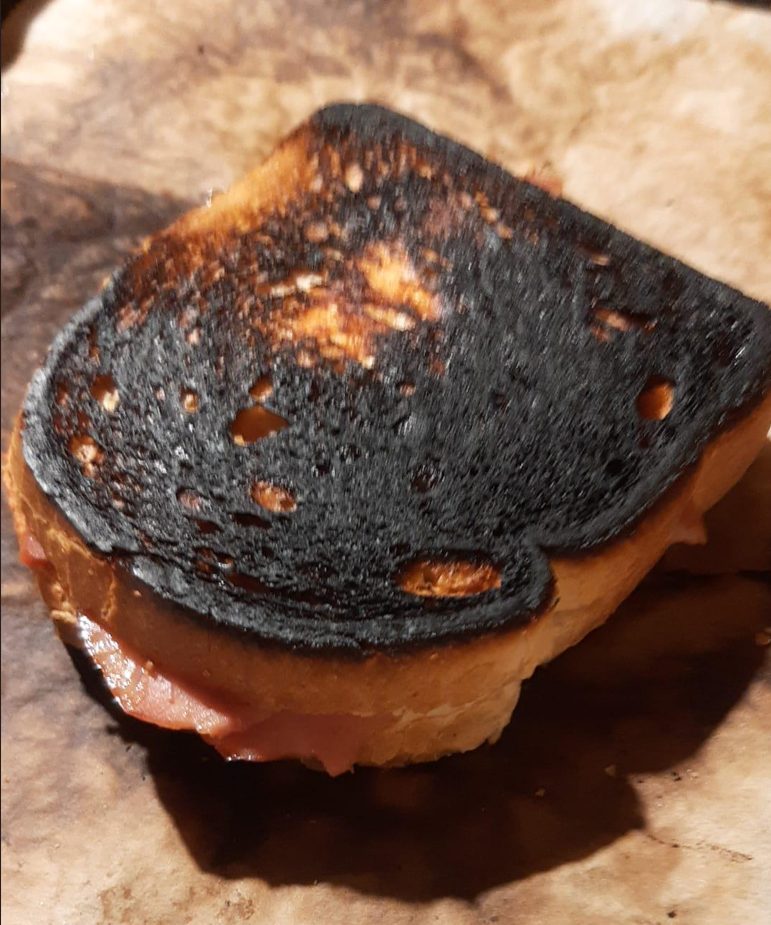 A close-up of a grilled sandwich with one side heavily burnt, resting on brown parchment paper—truly the epitome of hilarious cooking disasters. The bread appears charred and blackened, with some filling visible at the edges.