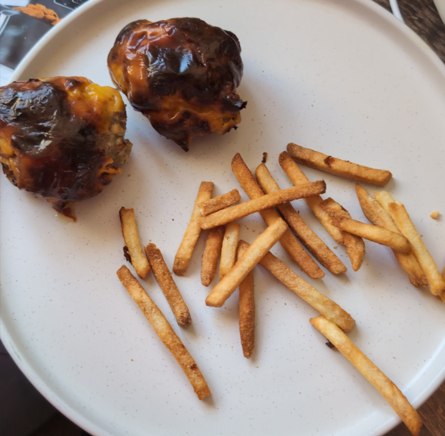 Two baked, cheese-topped meatballs with barbecue sauce on a white plate next to a scattered pile of French fries.