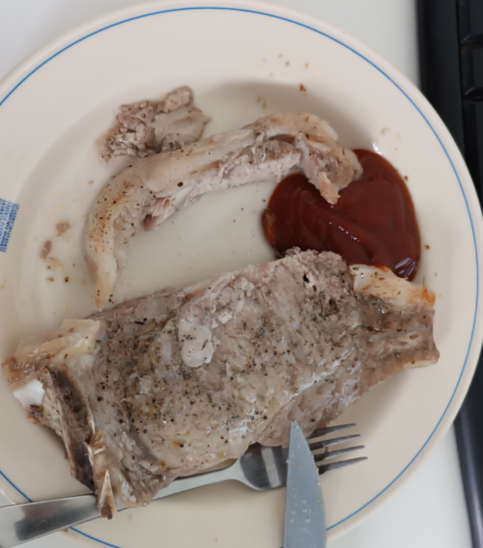 A partially eaten, seasoned piece of meat with some bones sits on a plate next to a portion of ketchup. A fork and knife rest on the plate’s edge.