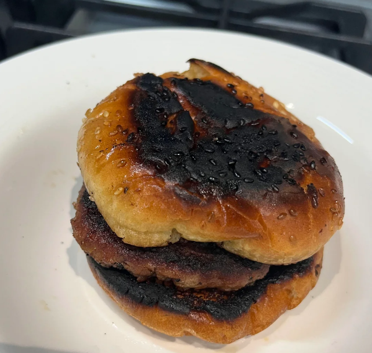 A burnt hamburger with a blackened bun and patty sits on a white plate, showing charred and overcooked surfaces.