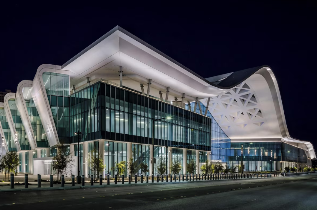 A modern, glass-walled convention center is brightly illuminated at night. Its striking white roof features bold curves and geometric patterns. The building stands along a quiet street lined with small trees.