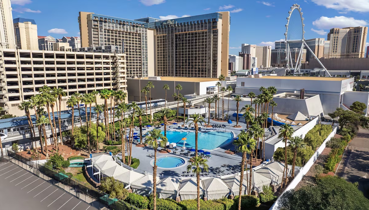 Aerial view of a hotel pool area with palm trees, lounge chairs, and hot tub, surrounded by tall buildings in Las Vegas. The High Roller Ferris wheel is visible in the background under a blue sky.