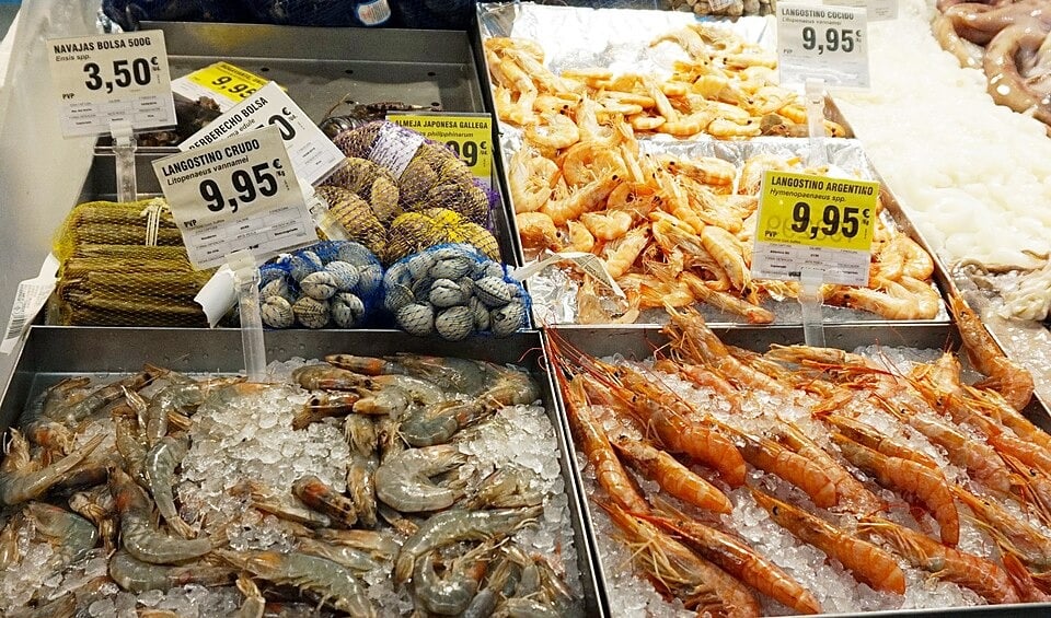 Fresh seafood at a market, including shrimp, prawns, clams, and mollusks displayed on ice, with labeled price tags above each section.