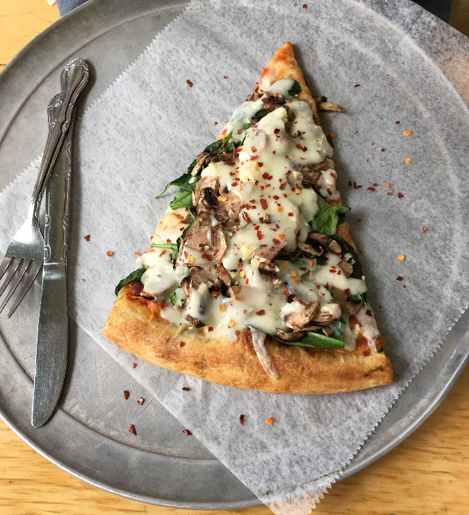 A quality slice of pizza for cheap, topped with mushrooms, spinach, melted cheese, and red pepper flakes, sits on a round metal tray with a fork and knife beside it. The pizza rests on a sheet of parchment paper.