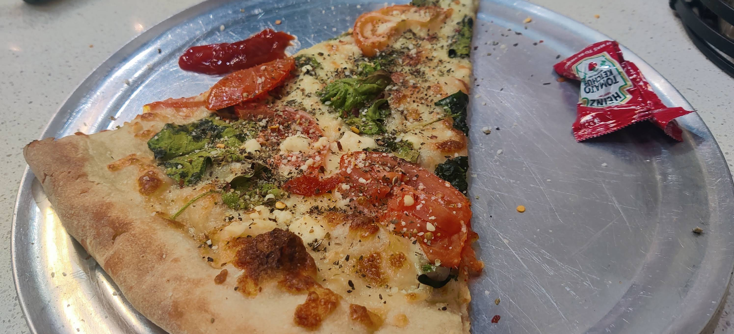 A quality slice of pizza for cheap, topped with tomatoes, broccoli, cheese, and herbs, rests on a metal tray. A small packet of crushed red pepper is open nearby, ready to add extra flavor.