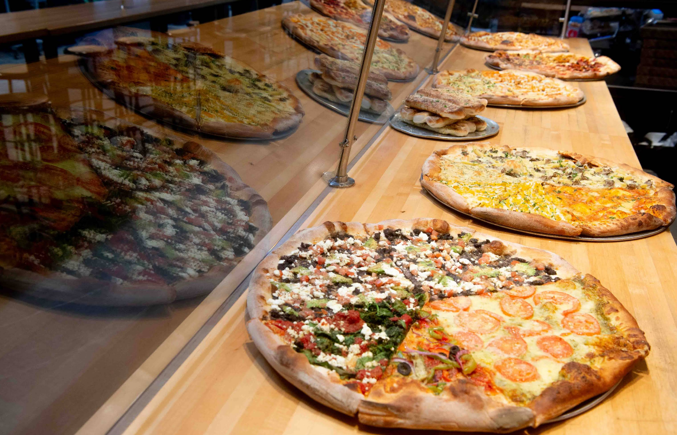 Several large pizzas with various toppings are displayed on a wooden counter behind a glass barrier, their reflections visible in the glass—a tempting view for anyone seeking a quality slice of pizza for cheap.