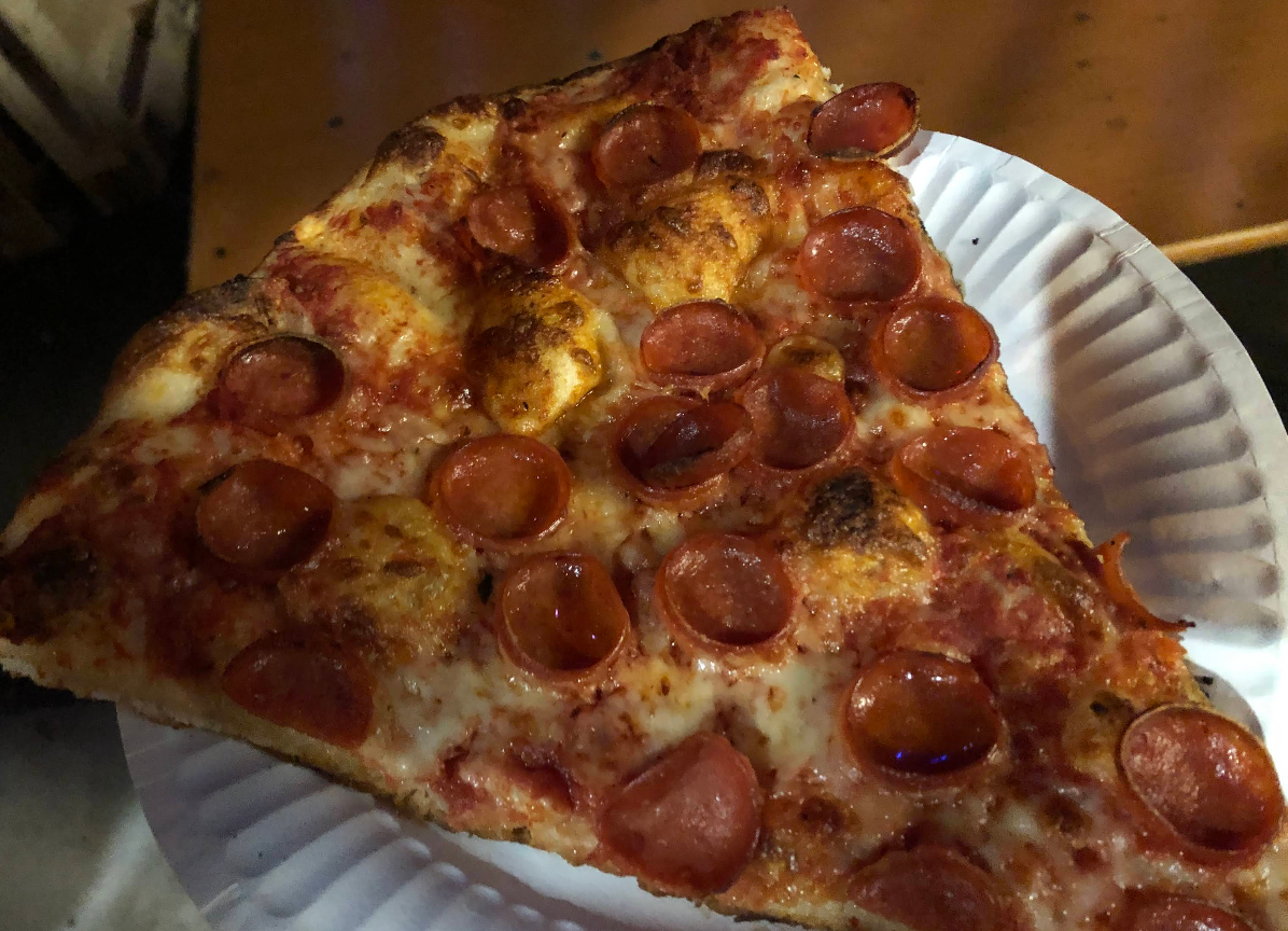 A large slice of pepperoni pizza with melted cheese and browned crust is served on a white, ridged paper plate—a quality slice of pizza for cheap, topped with plenty of pepperoni and a slightly crispy, chewy crust.