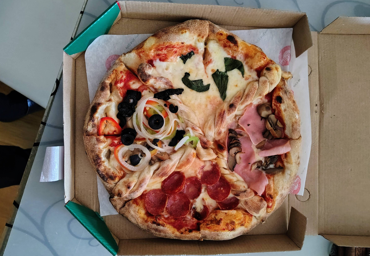 A pizza in an open box is divided into four quarters, each offering a quality slice of pizza for cheap—choose from pepperoni, ham and mushrooms, spinach, or mixed vegetables with olives, onions, and peppers.