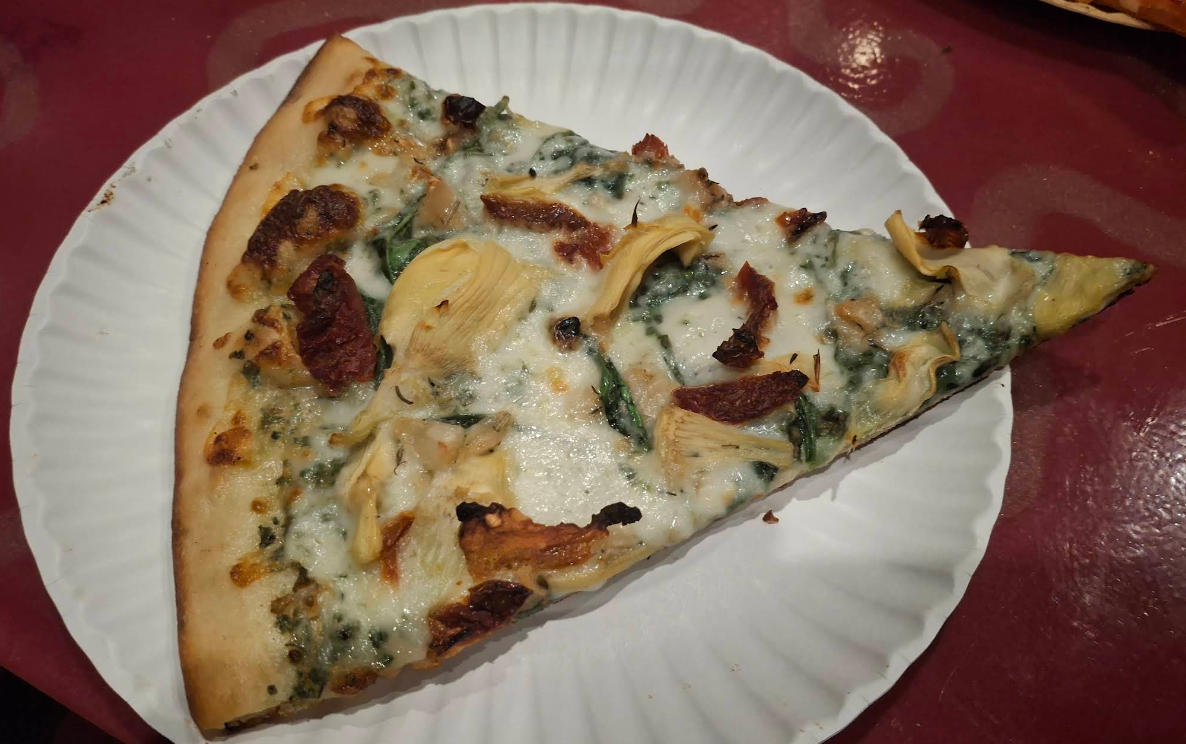 A quality slice of pizza for cheap, topped with spinach, artichoke, sun-dried tomatoes, and melted cheese, sits on a white paper plate atop a maroon tabletop.