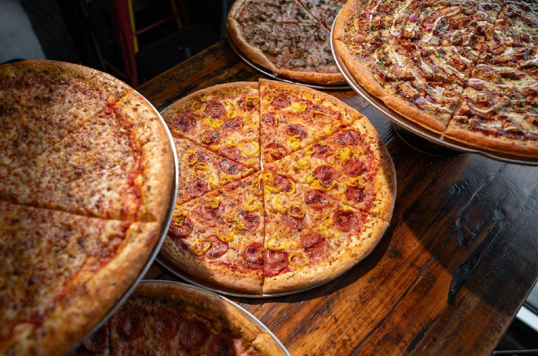 Four large pizzas on metal trays are displayed on a wooden table, each topped with pepperoni, peppers, sausage, and more—perfect for anyone craving a quality slice of pizza for cheap.