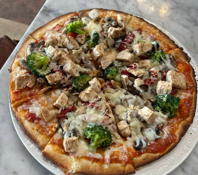 A quality slice of pizza for cheap on a white plate, topped with grilled chicken, broccoli florets, mushrooms, red bell peppers, and melted cheese, sits on a marble surface.