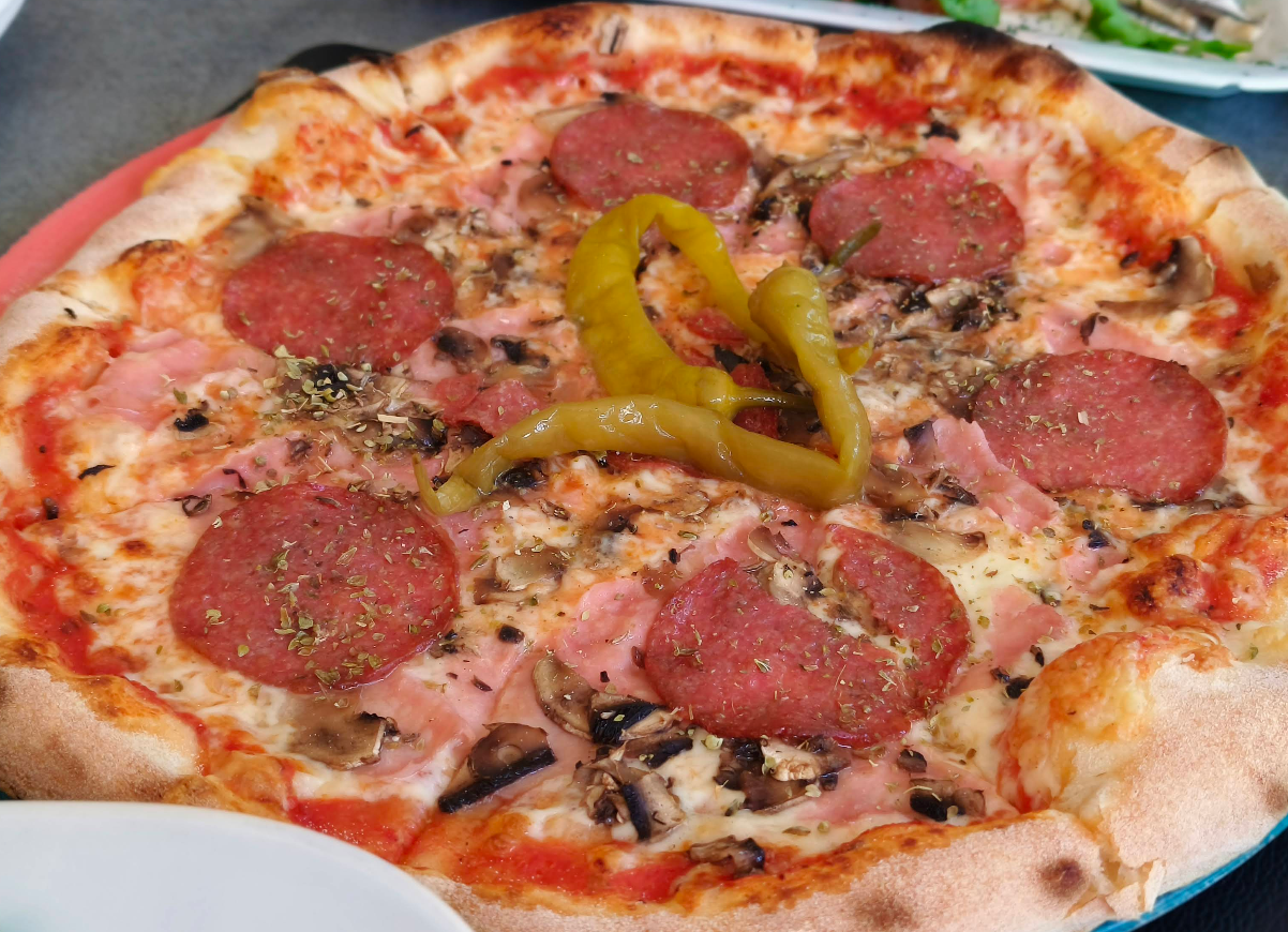 A close-up of a freshly baked pizza topped with pepperoni slices, mushrooms, diced ham, herbs, and two long yellow pepperoncini peppers in the center on a round plate—a quality slice of pizza for cheap.