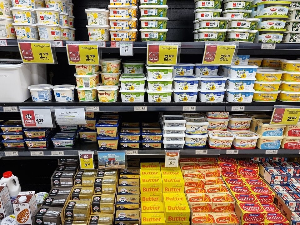 Grocery store shelves filled with various brands and types of butter and margarine in tubs and blocks, each labeled with prices, arranged in rows.