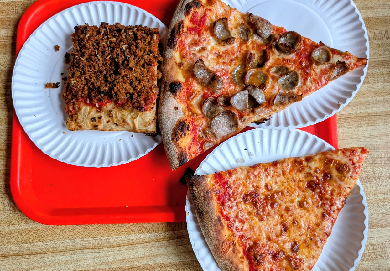 Three pizza slices on white paper plates sit on a red tray: one square slice with crumbled sausage, one large sausage slice, and one large plain cheese—perfect for anyone seeking a quality slice of pizza for cheap on a wooden table.