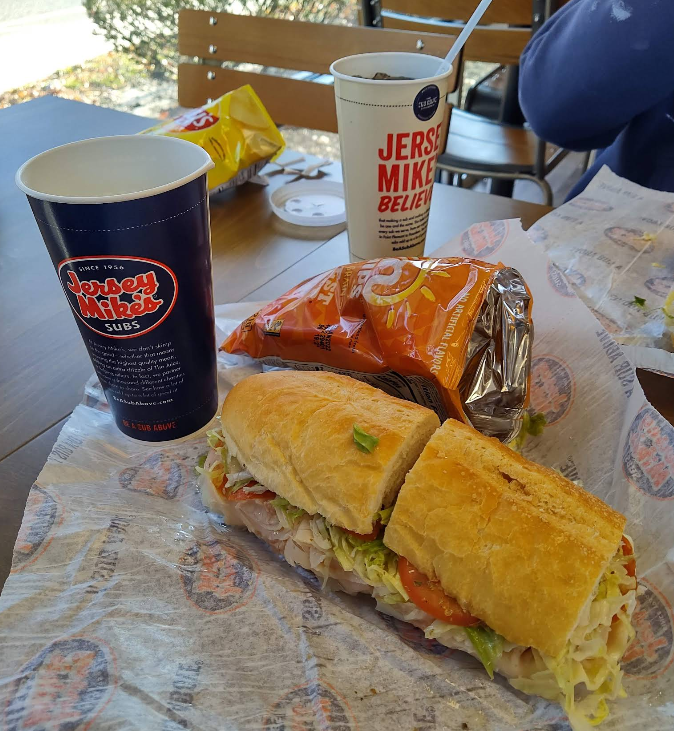 A Jersey Mike’s sub, from one of the top sandwich and sub chains, features turkey, lettuce, and tomato on branded paper. Nearby are a bag of chips, a signature blue drink cup, a white cup, and inviting outdoor seating.
