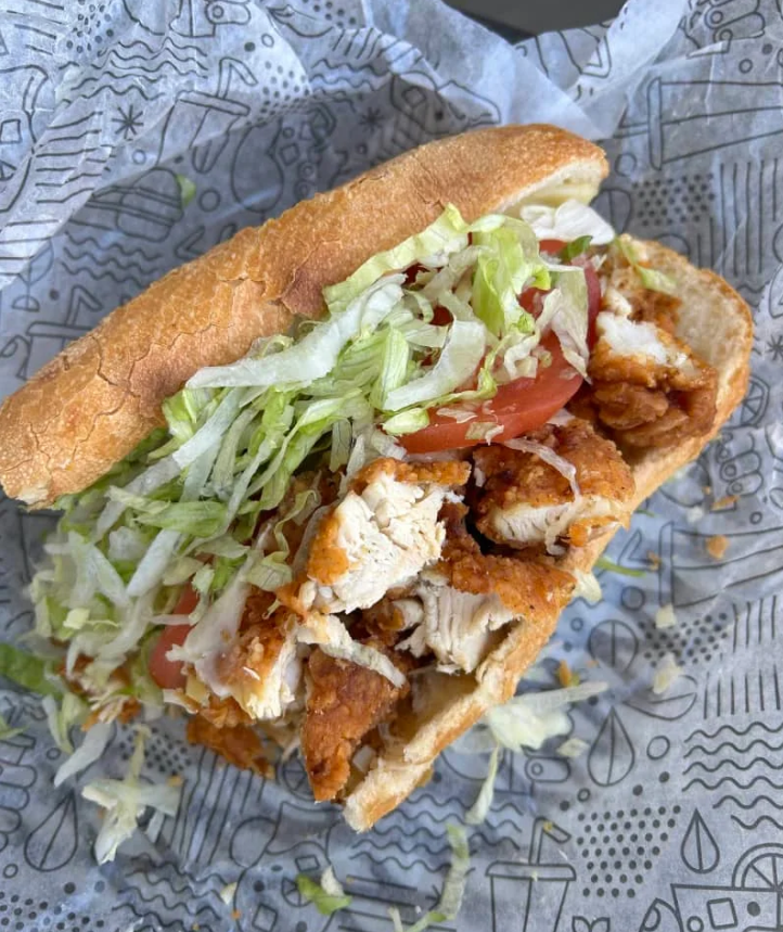 A close-up of a sandwich on patterned paper, filled with crispy fried chicken pieces, shredded lettuce, and tomato slices in a soft sub roll—just like those found at top sandwich and sub chains.