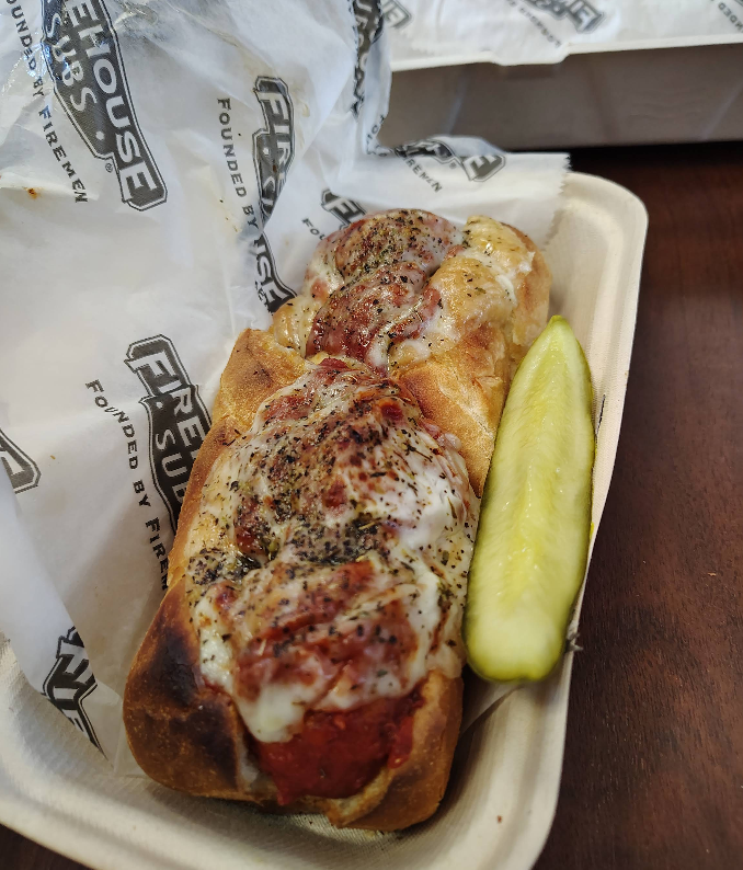 A toasted sub sandwich from one of the top sandwich and sub chains is topped with melted cheese, herbs, and tomato sauce in a takeout tray beside a dill pickle spear, all on Firehouse Subs branded paper.