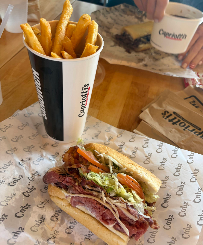 A cup filled with golden French fries sits next to a sub sandwich with roast beef, lettuce, tomatoes, and cheese on Capriotti’s-branded paper, showcasing why this spot stands out among top sandwich and sub chains. In the background, hands hold a cup and sandwich.