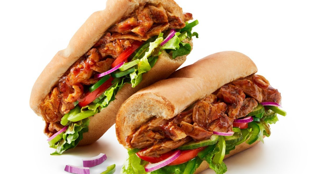 Two sub sandwiches, reminiscent of classic sandwich and sub chains, are filled with shredded barbecue meat, lettuce, tomato slices, green bell pepper strips, and red onion, set against a white background.