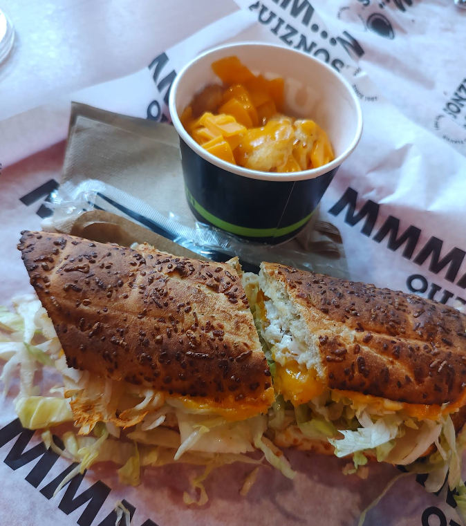 A toasted sandwich, inspired by popular sandwich and sub chains, is filled with lettuce, cheese, and chicken. It rests on branded paper next to a small cup containing a serving of diced mango.