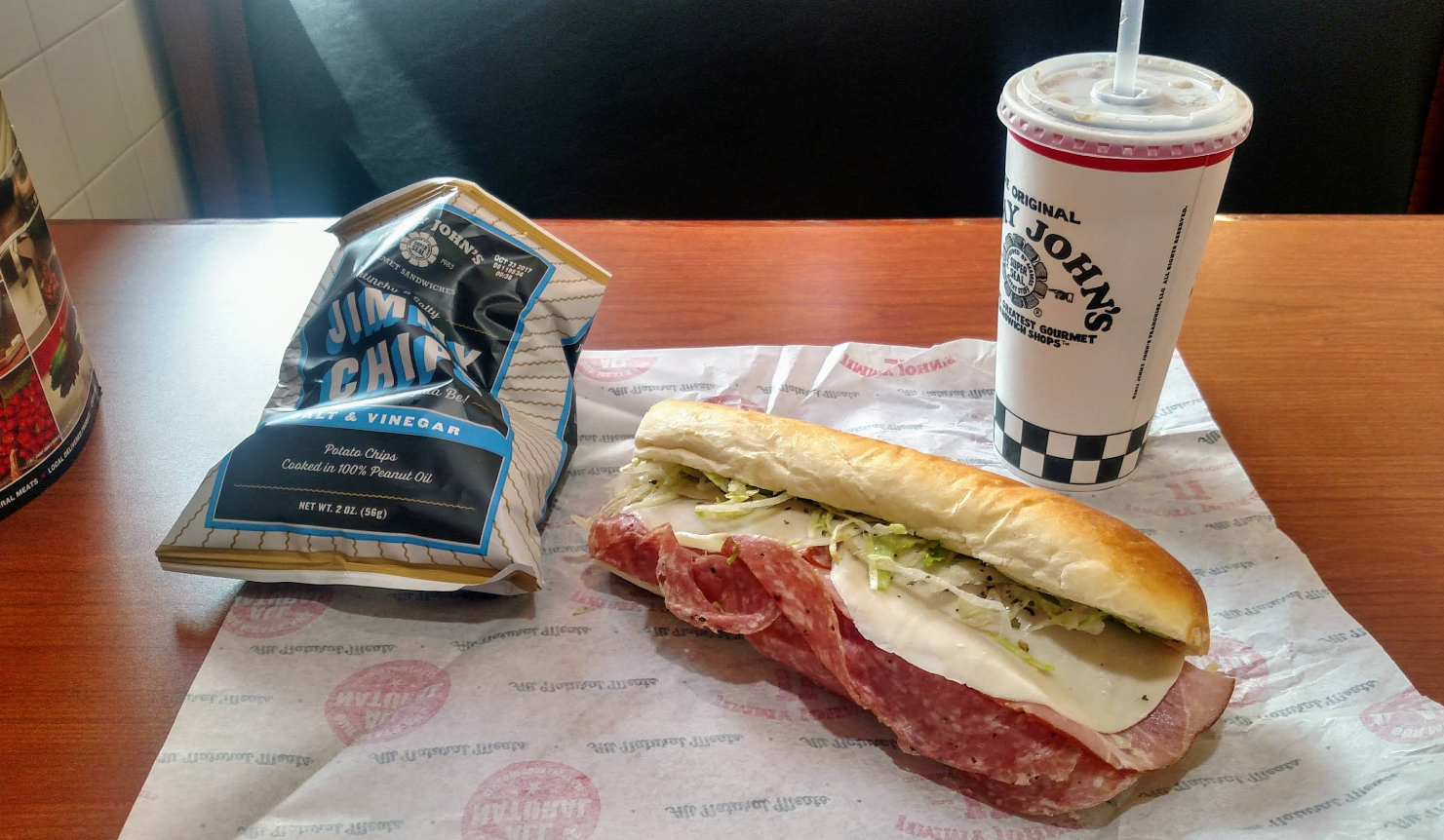 A sub sandwich from one of the top sandwich and sub chains sits on paper, next to a bag of Jimmy Chips and a large Jimmy John's drink on a wooden table.