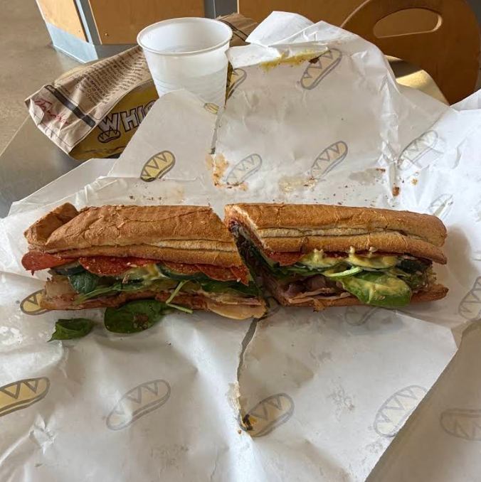 A sub sandwich from one of your favorite sandwich and sub chains sits cut in half on branded paper, filled with lettuce, spinach, avocado, meat, and cheese. A white plastic cup and brown paper bag are in the background.