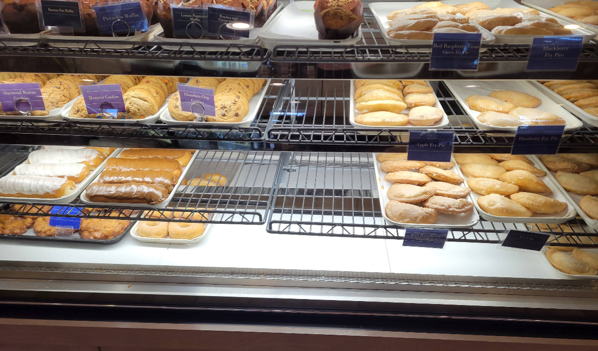 A bakery display case filled with assorted pastries and cookies—like muffins, chocolate chip cookies, turnovers, fruit pies, and cream-filled desserts—offers a tempting glimpse of an all-you-can-eat breakfast buffet arranged neatly on trays.