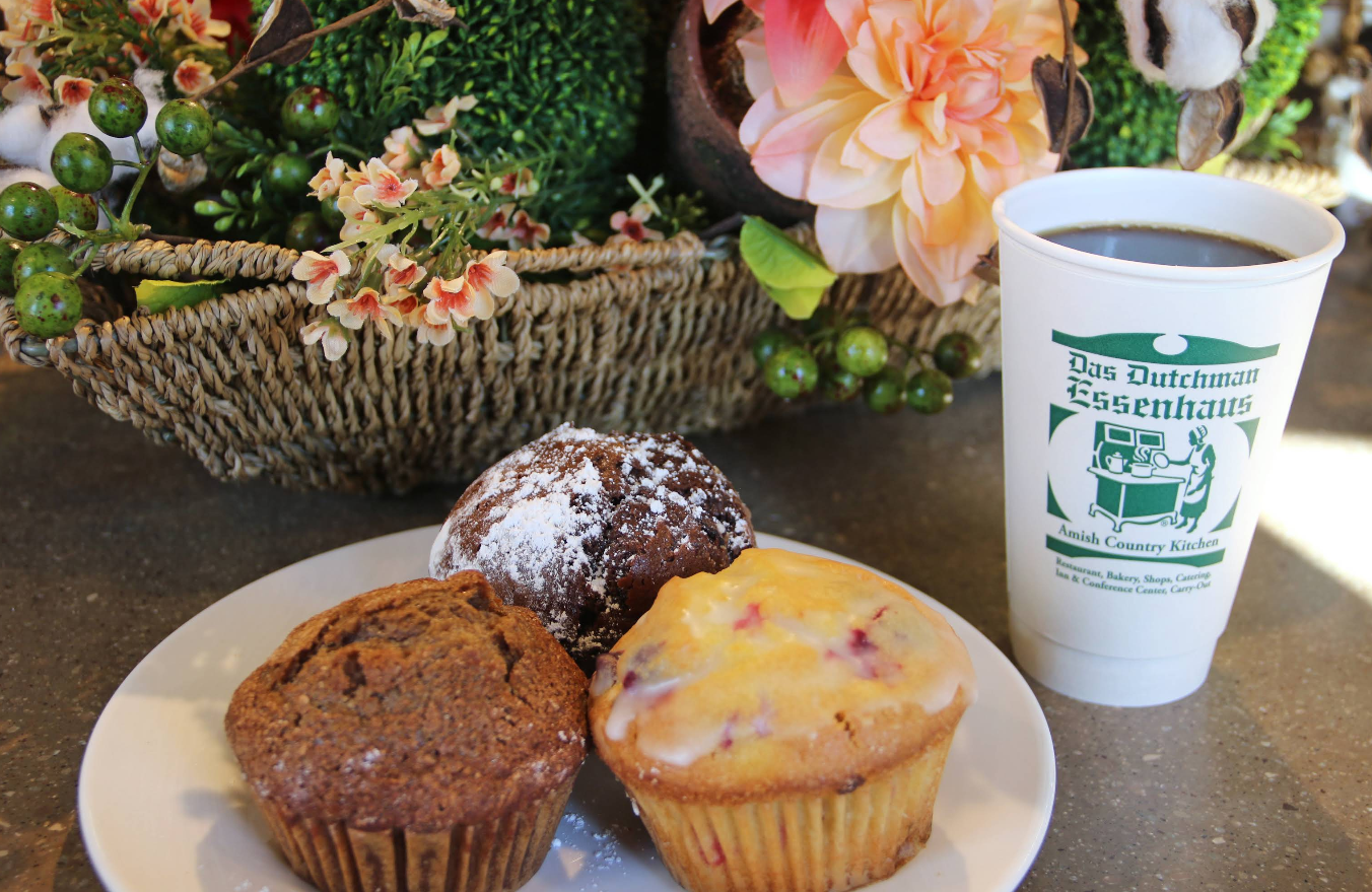 A plate with three muffins sits next to a cup of coffee in a white cup labeled "Das Dutchman Essenhaus," where an all-you-can-eat breakfast buffet awaits. A basket of artificial flowers and greenery is in the background.