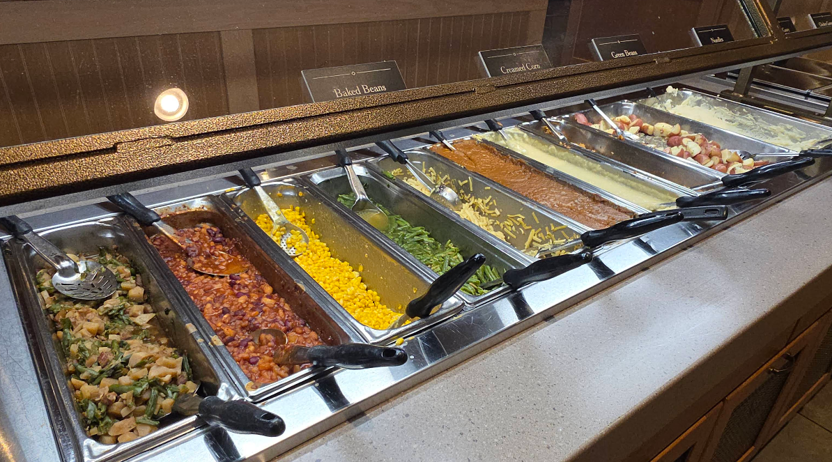 An all-you-can-eat breakfast buffet with trays of baked beans, corn, green beans, creamed corn, mashed potatoes, gravy, and diced potatoes, all lined up under a glass sneeze guard.