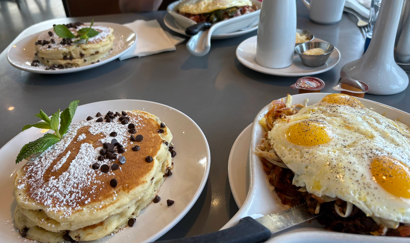 Plates of breakfast food on a table at an all-you-can-eat breakfast buffet: chocolate chip pancakes with powdered sugar and a mint sprig, plain pancakes, eggs sunny side up over hash browns, plus syrup, butter, and coffee cups.