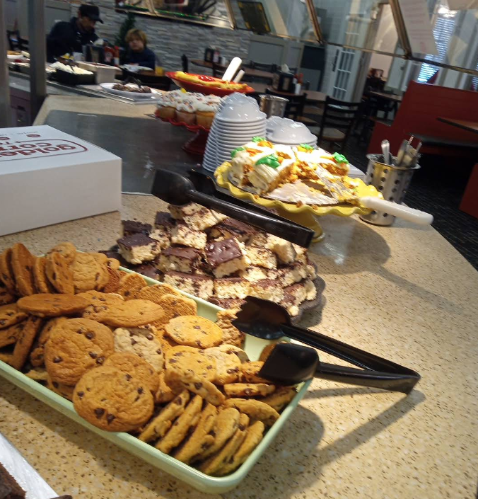 At an all-you-can-eat breakfast buffet, a dessert station offers trays of chocolate chip cookies, chocolate-topped bars, and assorted frosted cookies. Black tongs rest on the trays as people serve themselves in the background.