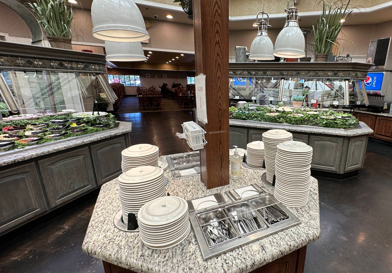 Stacks of plates and silverware are arranged on a granite counter in the foreground, with salad bars filled with vegetables and greens in the background inside a spacious all-you-can-eat breakfast buffet restaurant.