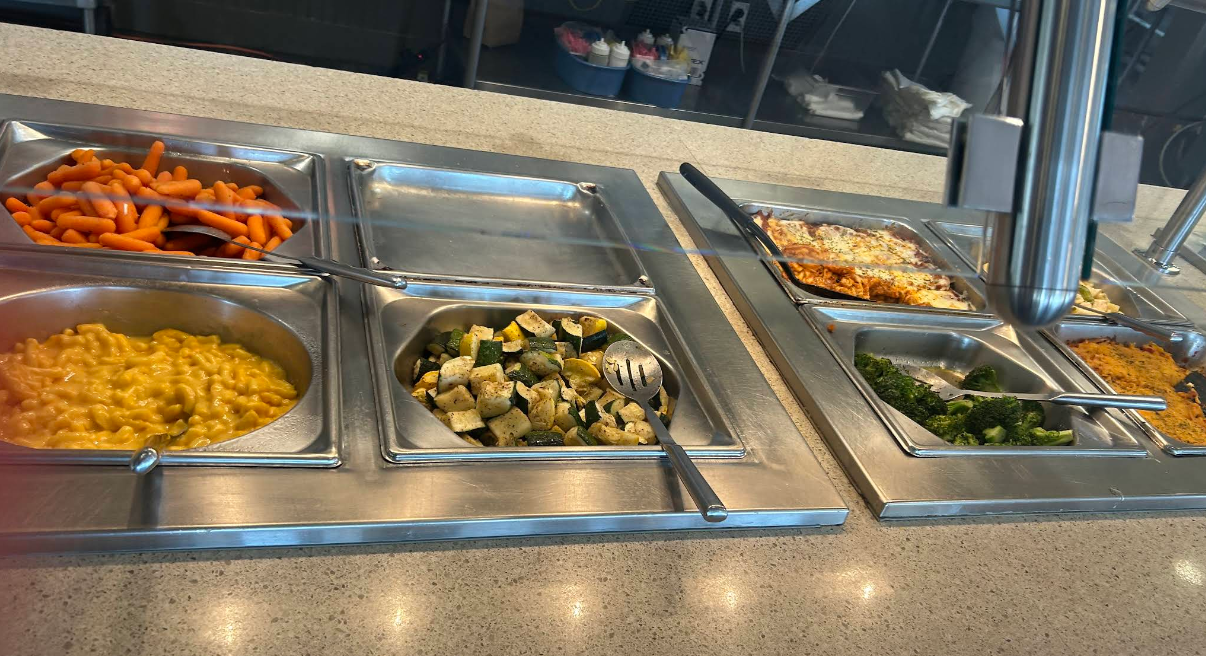 An all-you-can-eat breakfast buffet features trays of baby carrots, macaroni and cheese, roasted zucchini and squash, broccoli, lasagna, and a rice or grain dish, all neatly arranged on a countertop under a sneeze guard.