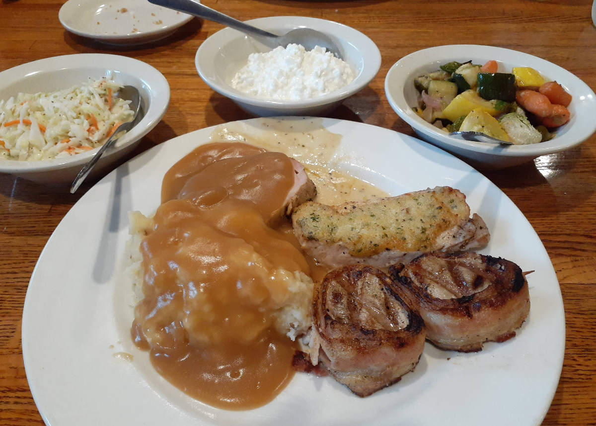 A plate with mashed potatoes and gravy, grilled meat, stuffed chicken, and two bacon-wrapped filets, served with bowls of coleslaw, cottage cheese, and mixed sautéed vegetables—perfect after an all-you-can-eat breakfast buffet on a wooden table.