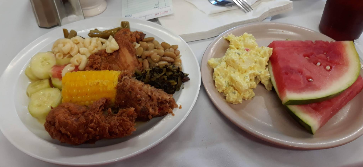 Two plates: one piled high like an all-you-can-eat breakfast buffet with fried chicken, macaroni, green beans, lima beans, collard greens, corn on the cob, cucumbers; another with potato salad and watermelon. A drink and utensils are in the background.