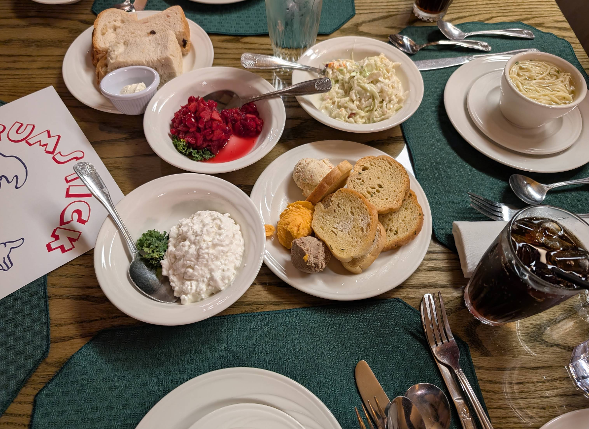A restaurant table set for an all-you-can-eat breakfast buffet, featuring plates of bread, cottage cheese, crackers with spreads, coleslaw, cranberry sauce, soup, a drink with ice, and utensils on green placemats.