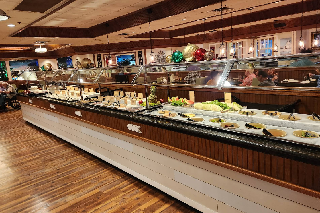 A long buffet counter in a restaurant displays an all-you-can-eat breakfast buffet with various salads, fruits, and sides under a glass sneeze guard. Diners are seated at tables in the background, with wood accents and decorative lighting enhancing the room.