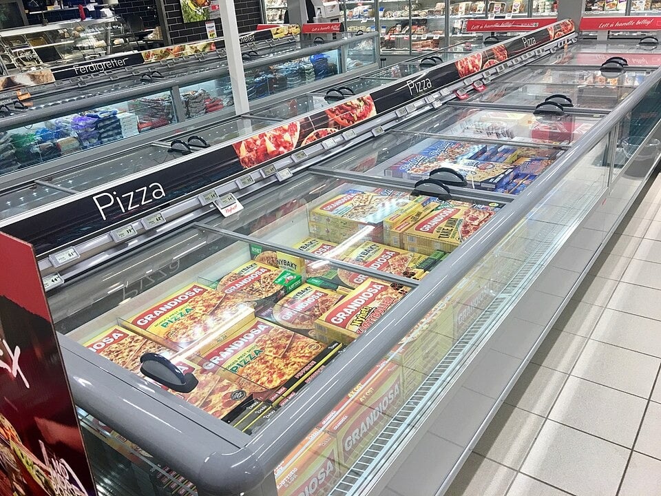 A supermarket freezer aisle filled with various brands and types of frozen pizzas in colorful boxes, organized behind glass doors and labeled "Pizza" above the section.