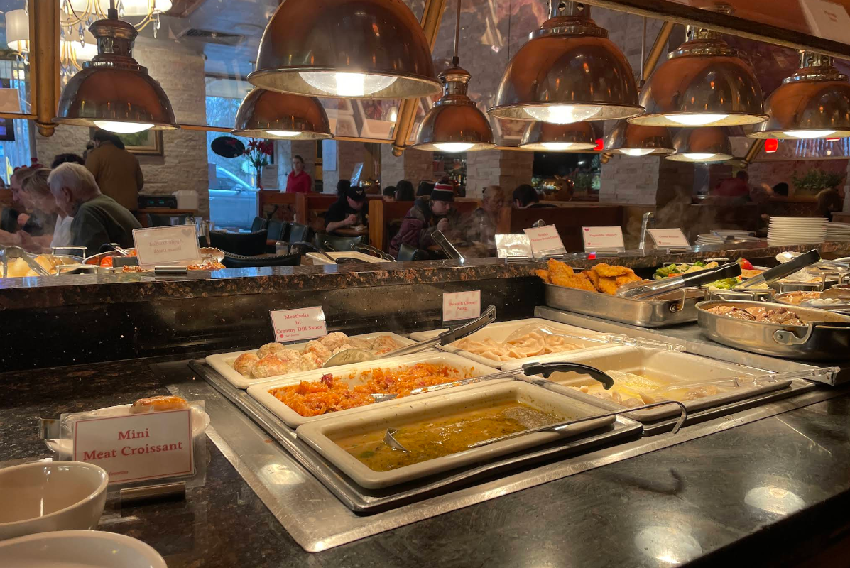 An all-you-can-eat breakfast buffet offers trays of hot food, including mini meat croissants, pasta dishes, and vegetables under warm lights, with people dining in the background.